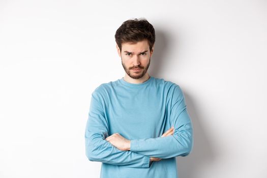 Image of offended and sad young man with beard, look from under forehead and sulking irritated, cross arms on chest defensive, mad at someone, standing on white background.