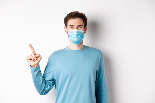 Coronavirus, health and quarantine concept. Caucasian man in medical mask pointing finger left, showing logo, standing in casual clothes on white background.