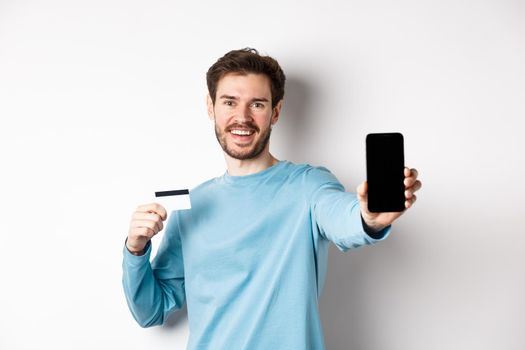 E-commerce and shopping concept. Smiling caucasian man showing plastic credit card and empty smartphone screen, recommending online app, white background.