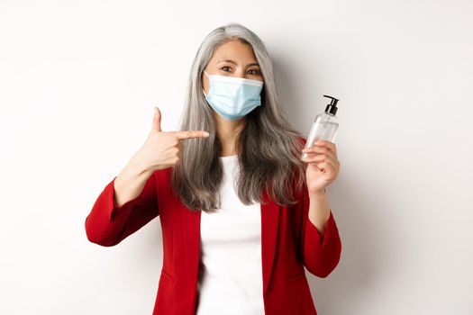 Covid, pandemic and business concept. Asian female manager in face mask, pointing finger at hand sanitizer, recommending antiseptic, white background.