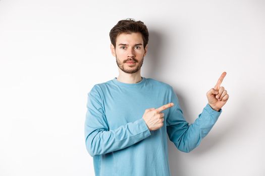 Portrait of serious caucasian man with beard pointing right at logo and looking at camera, standing in casual clothes on white background.