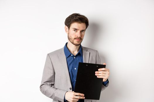 Young male ceo manager holding clipboard with documents and looking at camera, inspecting office, standing on white background.