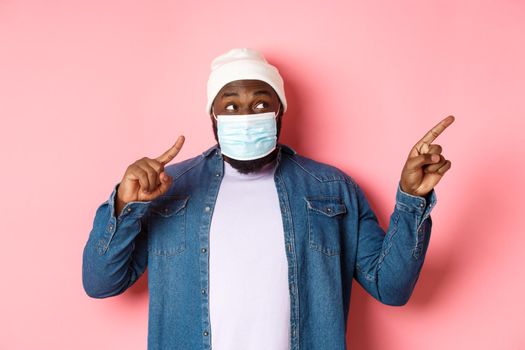 Covid-19, lifestyle and lockdown concept. Amused Black man in medical mask, looking and pointing right at copy space, standing over pink background.