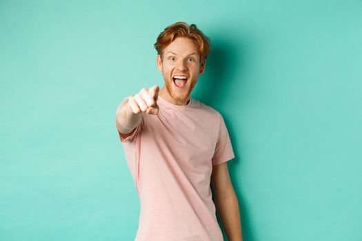Excited young man with red hair checking out something cool, pointing finger at camera and smiling fascinated, standing over turquoise background.