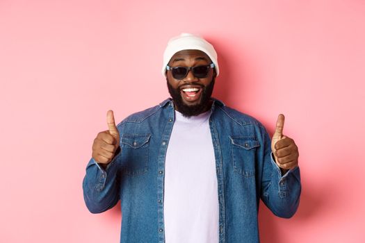 Satisfied african-american hipster guy in beanie and sunglasses showing thumbs-up, approve and praise good choice, smiling pleased, pink background.