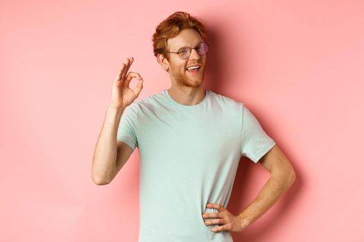 Cheerful guy with red hair and beard, wearing glasses, showing OK sign in approval and winking, smiling satisfied, standing over pink background.