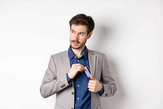 Confident and successful businessman looking self-assured while put plastic credit card in suit pocket, standing on white background.