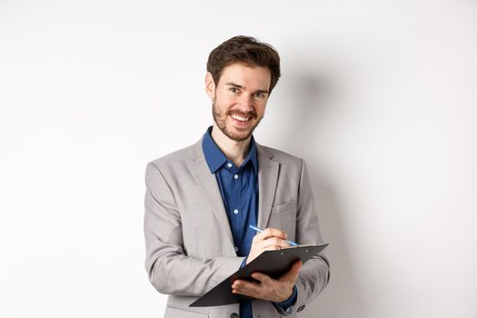 Smiling male manager in suit writing on clipboard, taking notes at office meeting, standing on white background.