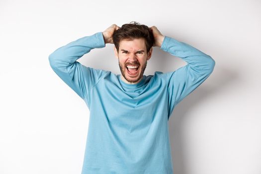 Mad young man pull hair oyt of head and screaming angry, shouting frustrated, standing hateful against white background.