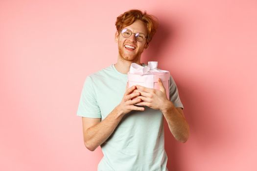 Valentines day and romance concept. Happy redhead boyfriend receive romantic gift, hugging box with present and saying thank you, smiling grateful, pink background.