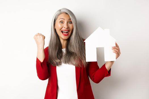 Happy asian grandmother showing paper house cutout and fist pump gesture, scream yes with joy, buying property, standing over white background.