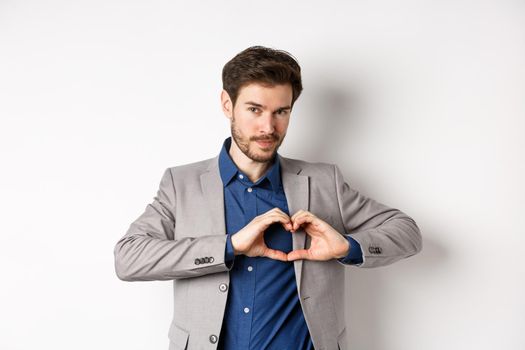 Romantic man in suit showing heart sign and smiling, love his girlfriend, standing on white background.