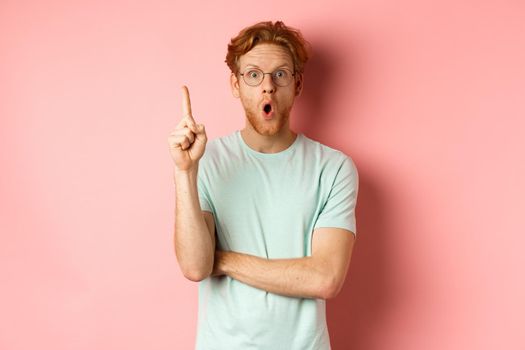 Attractive young man with red hair, raising finger in eureka gesture and pitching at idea, standing over pink background.