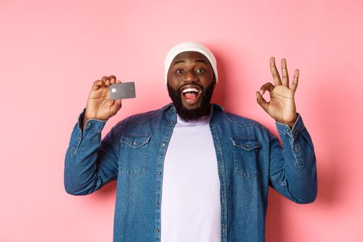 Shopping concept. Handsome african-american man recommending bank, showing credit card and okay sign, smiling satisfied, standing over pink background.