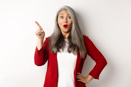 Impressed asian female entrepreneur in red blazer, pointing upper left corner and staring amazed at camera, checking out advertisement, white background.