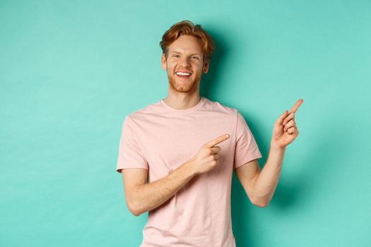 Handsome male model with red messy hair showing advertisement on copy space, pointing at upper right corner and smiling happy, standing over turquoise background.