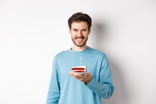 Celebration. Handsome young man celebrating birthday, holding bday cake with lit candle and smiling, making wish, standing over white background.
