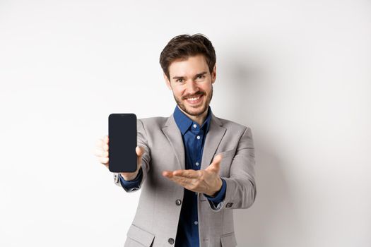 E-commerce and online shopping concept. Here you go. Smiling pleasant salesman demonstrate promo on phone, showing empty smartphone screen, white background.