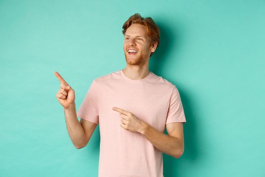 Dreamy handsome guy with red hair, smiling and looking at upper left corner with awe, admire something, standing over mint background. Copy space