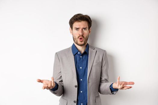 Confused and shocked businessman raising hands up and asking what happened, frowning disappointed, waiting for explanation, standing in suit on white background.