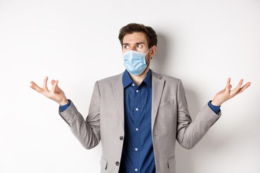 Confused businessman in medical mask shrugging and looking aside, know nothing, cant understand something, standing on white background.