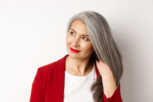 Beauty and haircare concept. Close up of elegant asian senior woman showing shiny and healthy grey hair, smiling and looking aside, white background.