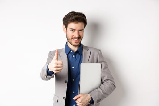 Successful businessman in stylish suit carry laptop and looking confident at camera, showing thumbs up in approval, white background.