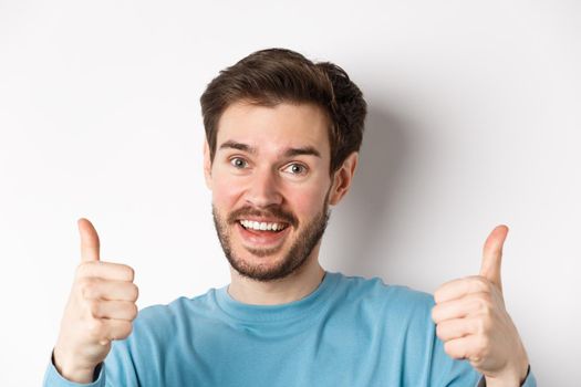 Close-up of cheerful man say yes, showing thumbs up in approval, praise good job, smiling approvingly, standing on white background.