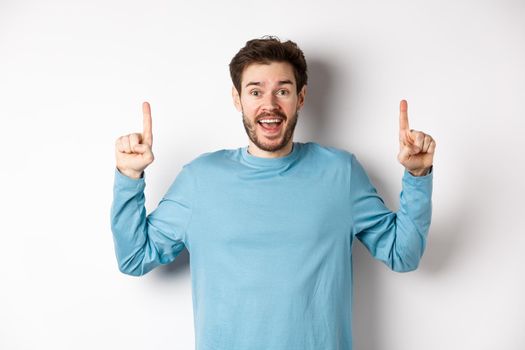 Cheerful young man showing advertisement with happy smile, pointing fingers up at awesome logo banner, standing on white background.