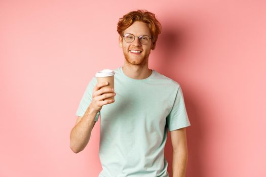 Happy redhead man in glasses and t-shirt drinking coffee and smiling, enjoying lunch break, holding takeaway cup, standing over pink background.