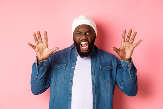 Angry african-american guy in beanie, scare you, roaring and screaming, showing hands, standing ove rpink background.