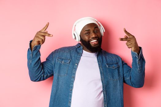 Cool african-american man dancing hip-hop, listening music in headphones, standing over pink background.