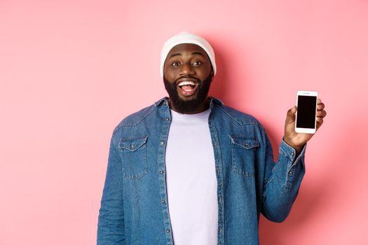 Handsome hipster guy in beanie and denim shirt smiling, showing mobile phone screen with happy face, recommend app, standing over pink background.