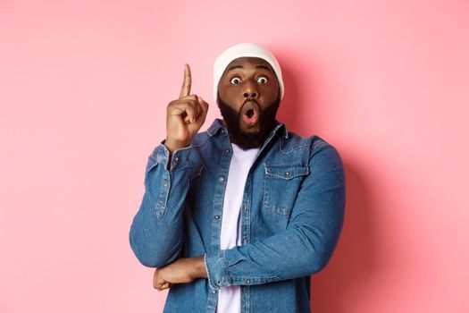 Handsome hipster Black guy having an idea, raising finger up and saying suggestion, got solution, standing over pink background.