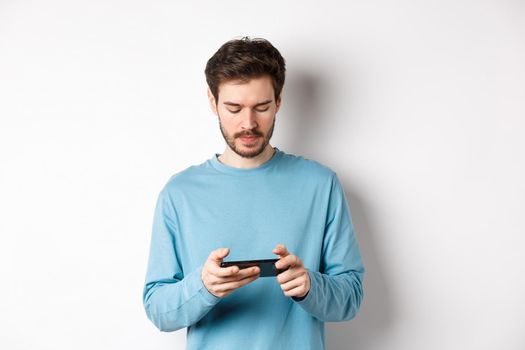 Male model playing video game on smartphone, looking serious at mobile screen, standing in sweatshirt over white background.