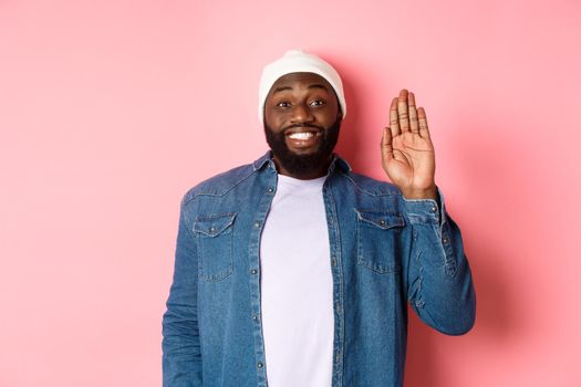 Friendly smiling Black man saying hello, waving hand, greeting you, standing over pink background.