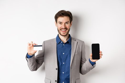 Online shopping. Smiling business man in suit showing plastic credit card with empty smartphone screen, standing against white background.