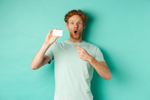 Shopping concept. Handsome redhead man in t-shirt showing plastic credit card and smiling, standing over turquoise background.