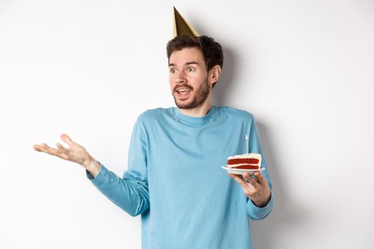 Celebration and holidays concept. Surprised man shrugging in disbelief, looking away, holding birthday cake and wearing party cone hat, white background.