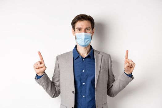 Covid-19, pandemic and business concept. Happy businessman in medical mask and suit pointing fingers up, standing on white background.