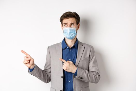 Covid-19, pandemic and business concept. Hesitant businessman in medical mask and suit looking, pointing left with doubtful face, standing on white background.