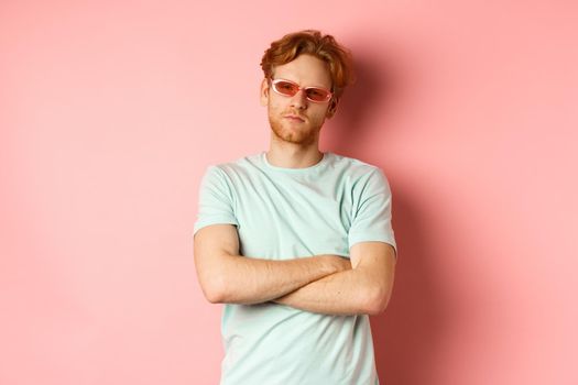 Tourism and vacation concept. Sassy redhead guy in sunglasses, cross arms on chest and looking serious at camera, standing over pink background.