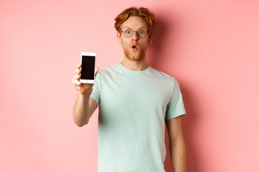Image of handsome guy with red hair, wearing glasses and t-shirt, saying wow and showing smartphone screen, standing against pink background.
