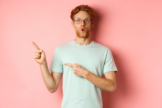 Shopping concept. Portrait of man with red hair and beard, wearing glasses with summer t-shirt, pointing fingers at upper left corner and saying wow impressed, pink background.