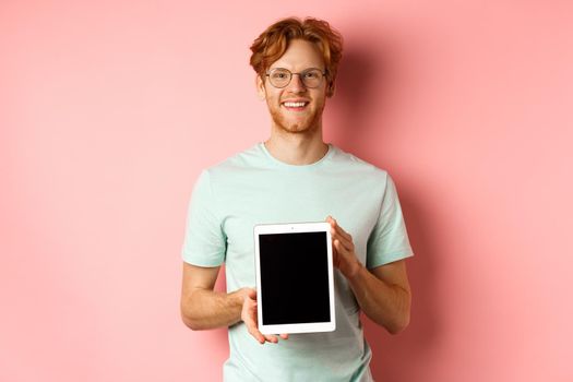 Funny redhead guy in glasses demonstrate digital tablet screen, smiling at camera, standing over pink background.