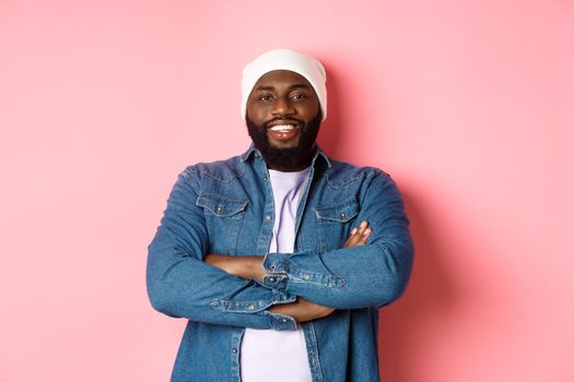 Handsome hip hop style Black man in beanie and denim shirt, smiling confident, cross arms on chest and staring at camera on pink background.