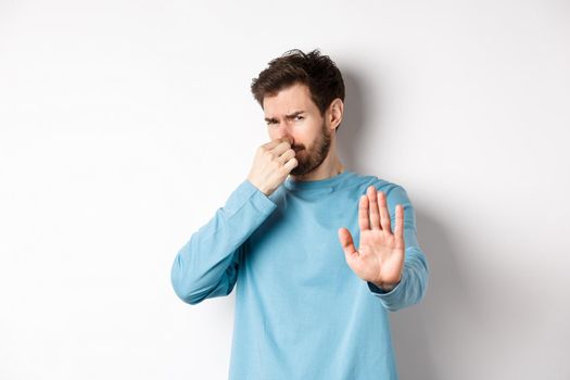 Image of caucasian man stretch out hands to stop or reject something, shut nose disgust, refusing product with bad smell, standing over white background.