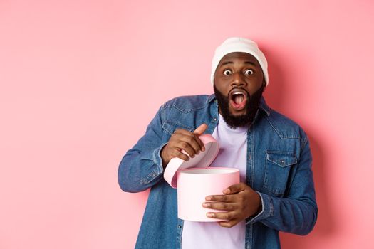 Excited Black man open box with gift and staring at camera grateful and amazed, receiving present, standing over pink background.