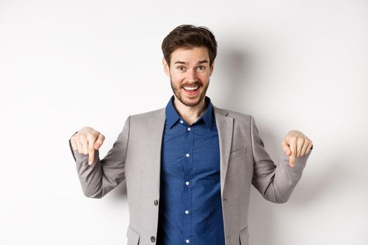 Hey check this out. Happy businessman in suit pointing fingers down and smiling, inviting to event, showing link, standing on white background.