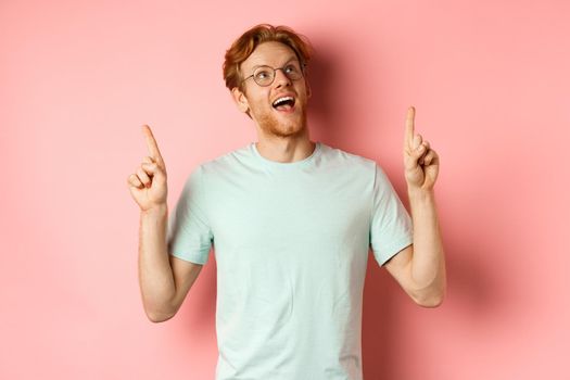 Amazed redhead guy in glasses checking out special deal, looking and pointing fingers up with amazed face, standing over pink background.
